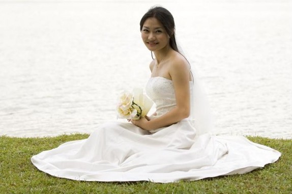A beautiful young woman sits on a grassy bank in her wedding dress